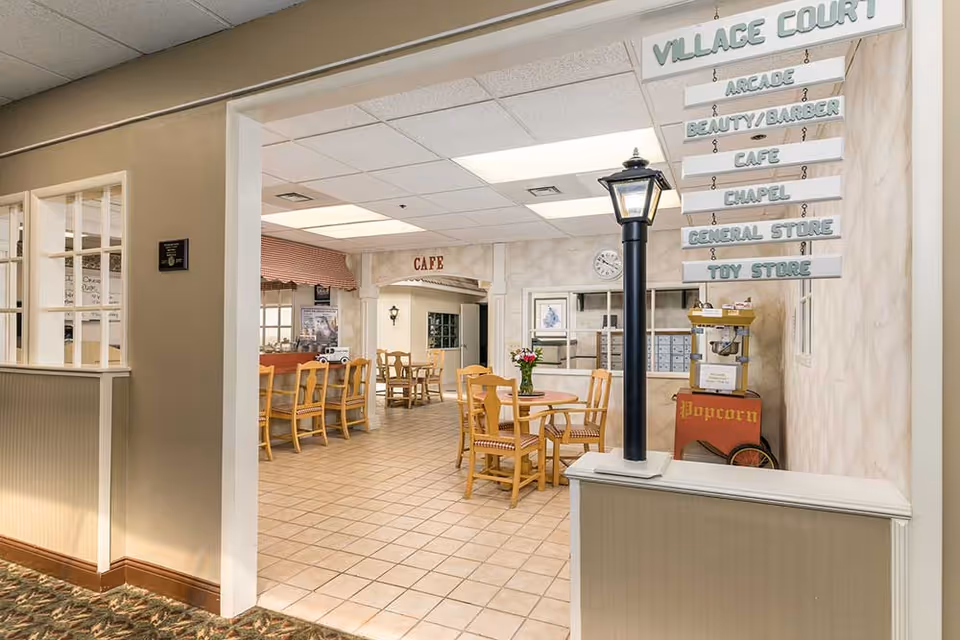 Interior view of a senior living facility common area with a small cafe. The space features wooden tables and chairs, a popcorn machine, and a decorative street lamp post. Hanging signs indicate directions to various amenities including Village Court, Arcade, Beauty/Barber, Cafe, Chapel, General Store, and Toy Store. The floor is tiled and the walls are light-colored with a textured finish.