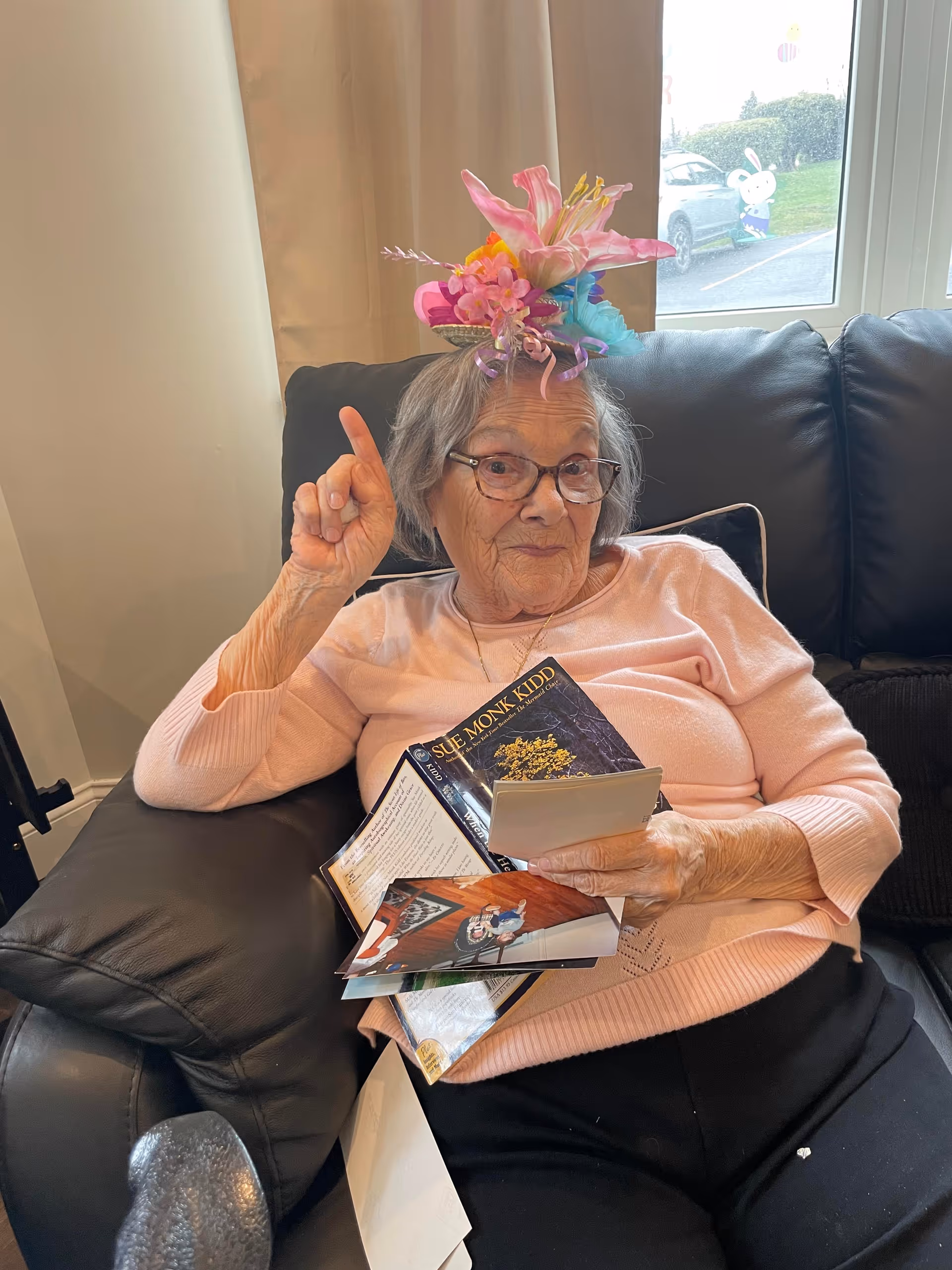 An older woman wearing glasses and a colorful floral headpiece sits on a black couch holding books and cards and raises one finger.
