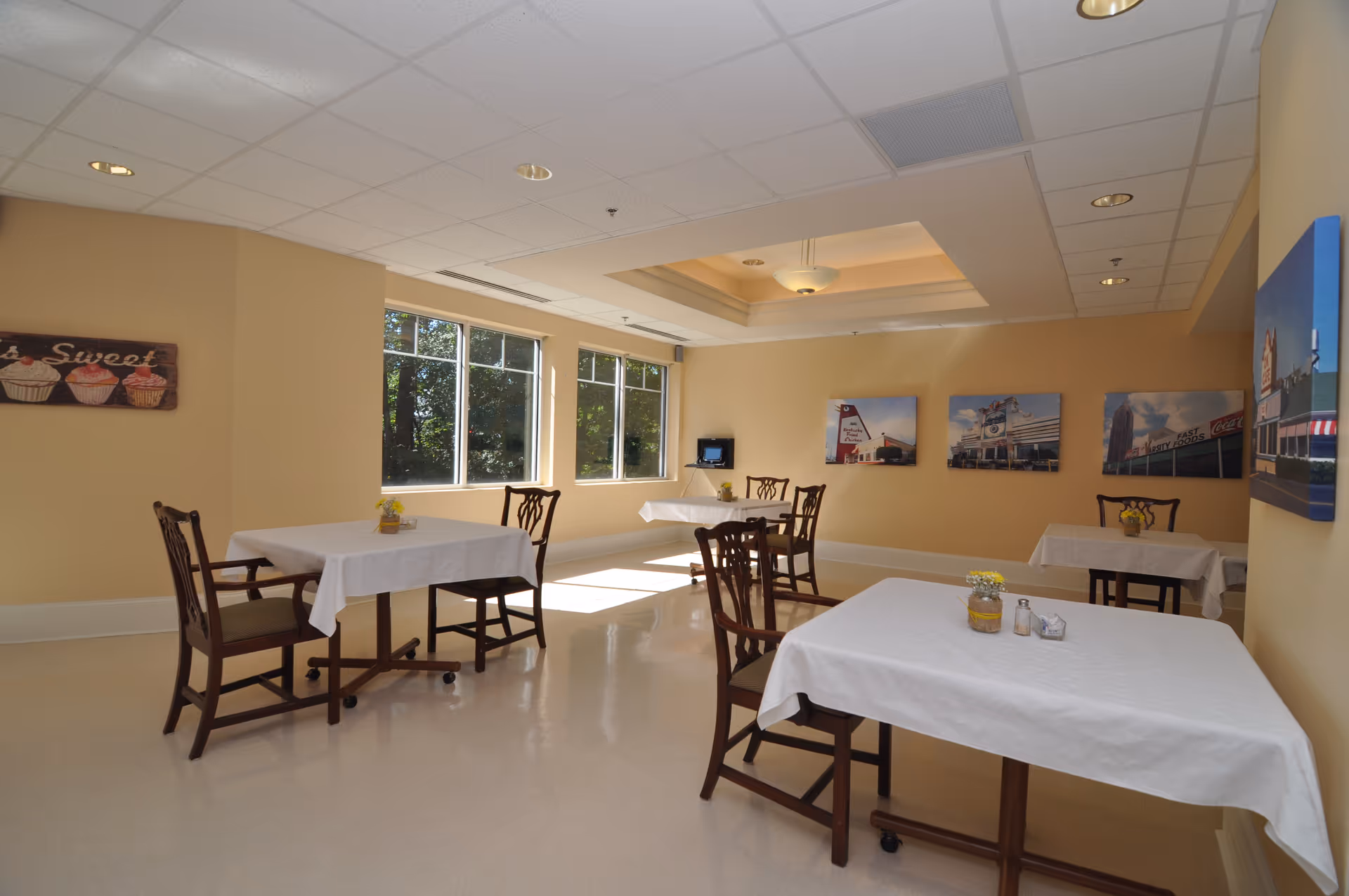 A bright dining room with several tables covered in white tablecloths, each accompanied by wooden chairs. The room has large windows letting in natural light, beige walls adorned with framed pictures, and a ceiling light fixture.