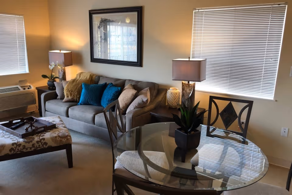 A cozy living room area with a gray sofa adorned with multiple cushions in blue and beige tones, a patterned ottoman with a wooden tray on top, two side tables each with a lamp, a glass-top round dining table with four chairs, and two windows with blinds partially closed.