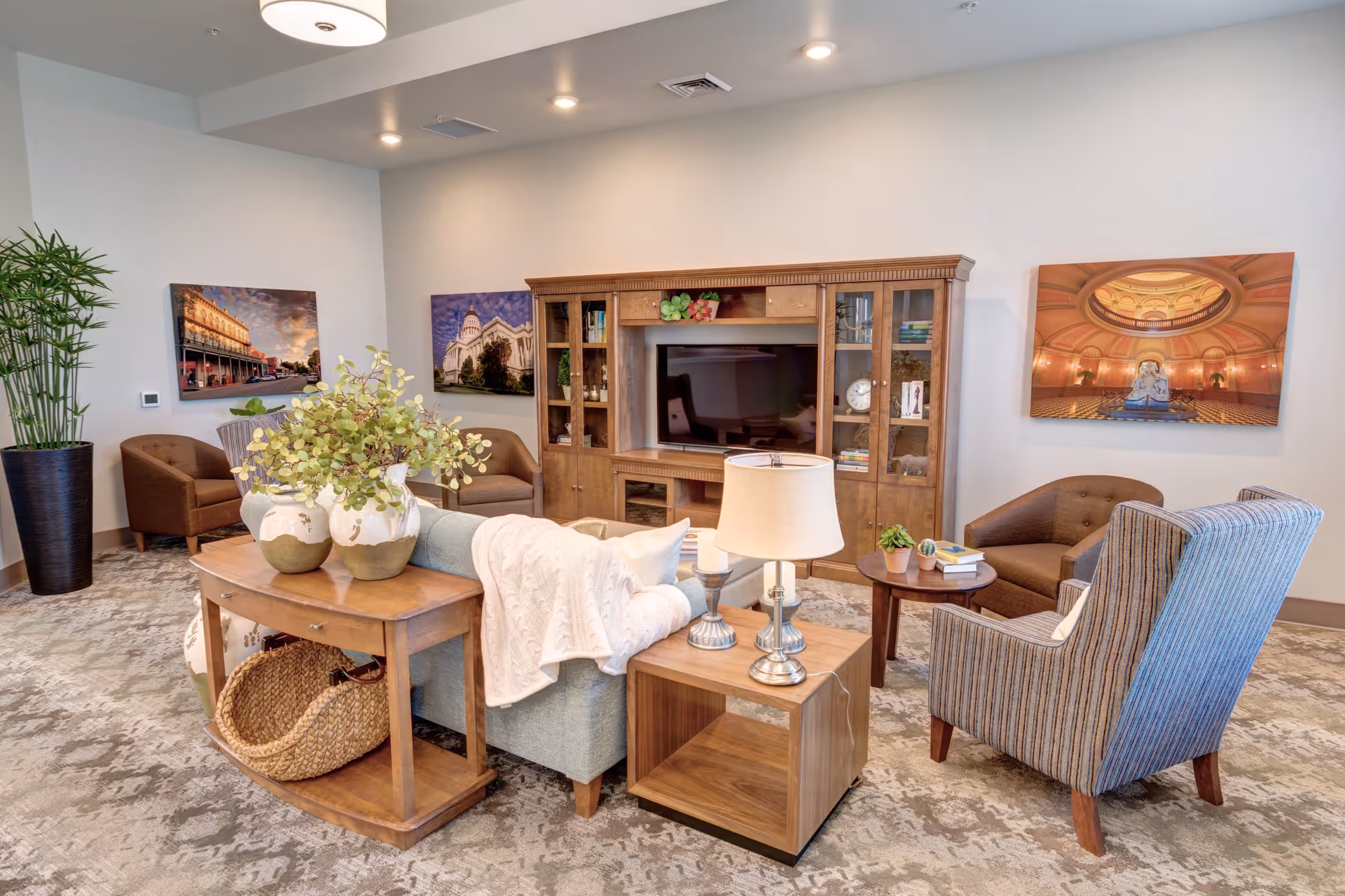 A cozy living room area with a wooden entertainment center housing a flat-screen TV, surrounded by various chairs including a striped armchair and brown cushioned chairs. A light blue sofa with a white throw blanket faces the TV. A wooden console table behind the sofa holds two decorative vases with greenery, and a small wooden side table with a lamp and small plants is next to the sofa. The room has a patterned carpet and three framed pictures on the walls, along with a tall potted plant in the corner.