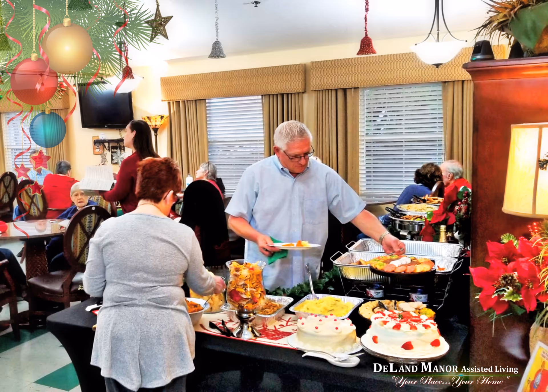 A festive dining area in an assisted living facility decorated for the holidays. Several elderly residents are seated at tables in the background, while two people serve themselves food from a buffet table filled with various dishes and desserts. Holiday decorations, including ornaments and poinsettias, are visible around the room.