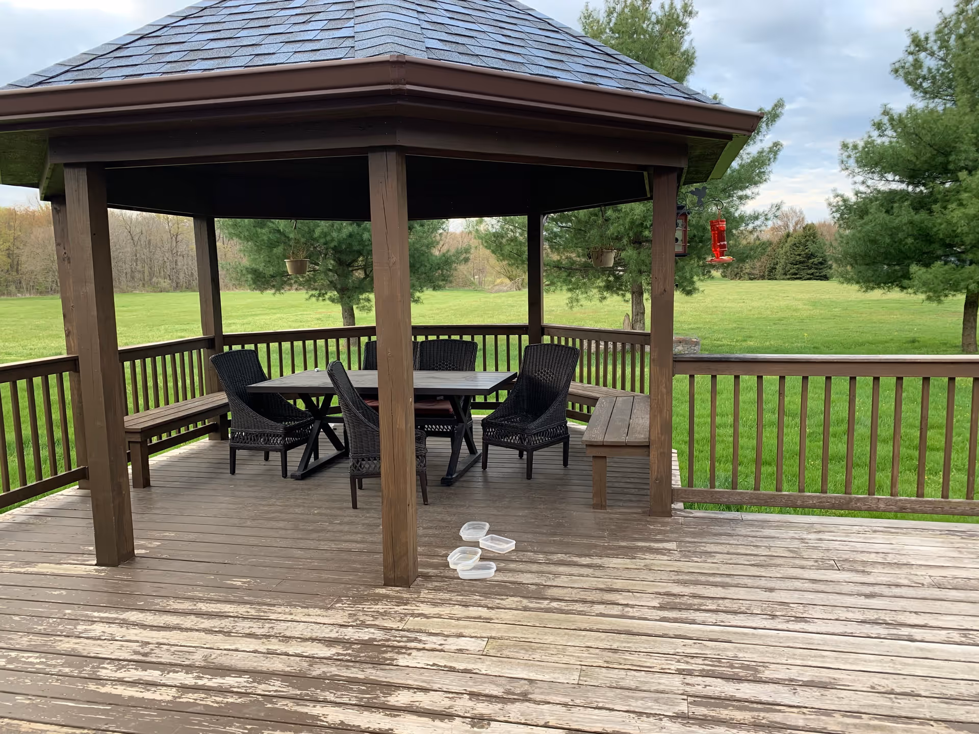 A wooden gazebo with a shingled roof situated on a wooden deck. Inside the gazebo, there is a rectangular table surrounded by six black wicker chairs. Two wooden benches are attached to the gazebo railing. The gazebo overlooks a large grassy field with trees in the background. Three small plastic containers are placed on the deck floor outside the gazebo.