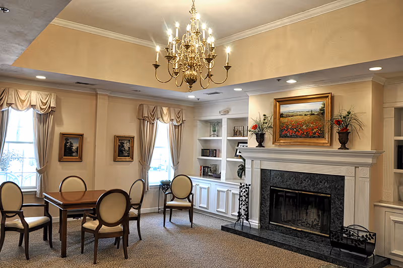 A cozy living room area with a wooden table and four upholstered chairs. The room features a large fireplace with a black marble surround and a painting of a field of flowers above it. Built-in white shelves with books and decorative items flank the fireplace. Two windows with beige curtains allow natural light to enter the room. A gold chandelier hangs from the ceiling, illuminating the space.