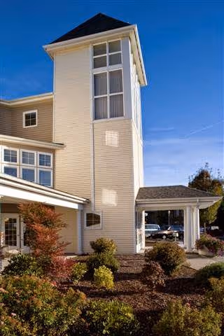 Exterior view of a multi-story building with beige siding and large windows under a clear blue sky. The building is surrounded by landscaped bushes and plants, with a covered entrance visible on the right side.