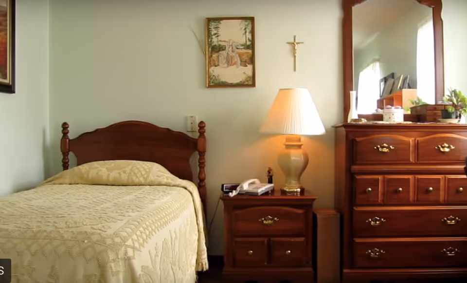 A cozy bedroom with a single wooden bed covered with a cream-colored quilt. Next to the bed is a wooden nightstand with a lamp, a telephone, and a small figurine. On the right side, there is a wooden dresser with a large mirror, decorated with small plants and other items. A framed picture and a crucifix hang on the wall above the nightstand.