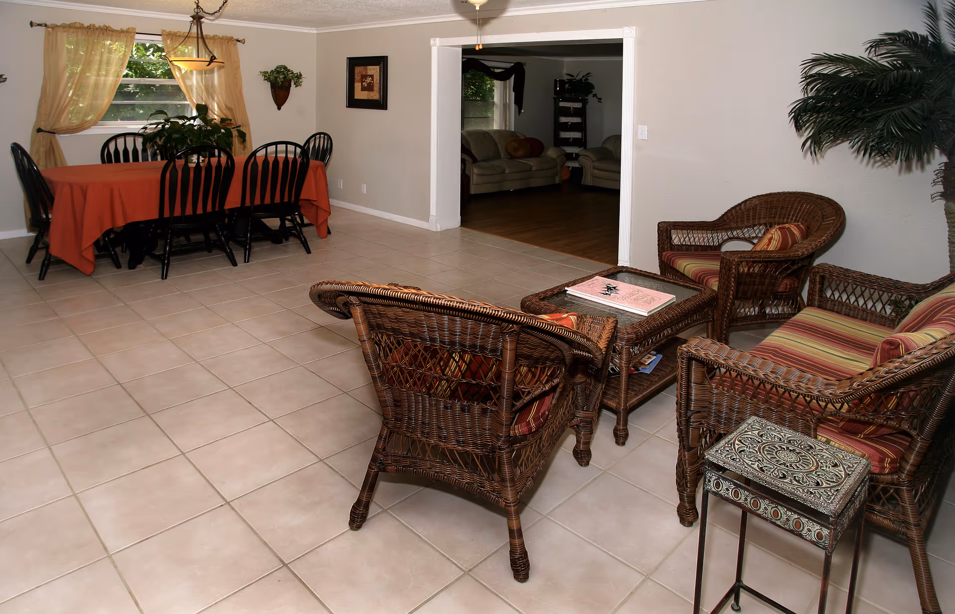 Interior view of a senior living facility showing a tiled floor with a wicker seating area consisting of three chairs and a coffee table with a book on it. In the background, there is a dining area with a table covered by a red tablecloth and six black chairs. The room has light-colored walls, a window with sheer curtains, and some plants for decoration.