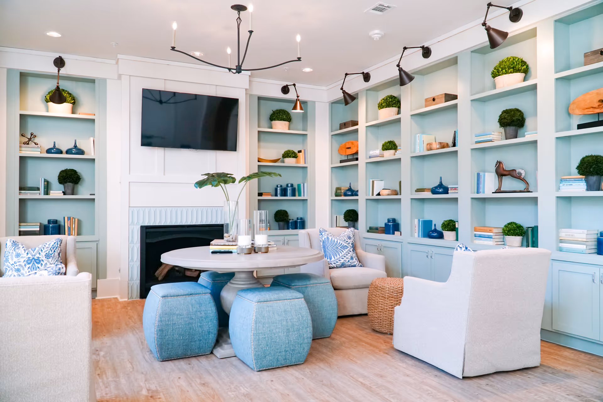 A cozy living room with light blue built-in shelves filled with books, plants, and decorative items. The room features a round wooden table surrounded by four blue cushioned stools and three beige armchairs with blue patterned pillows. A flat-screen TV is mounted above a fireplace, and the room is lit by ceiling lights and wall-mounted spotlights.