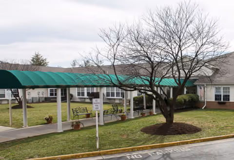 Outdoor view of Cardinal Senior Living at Roanoke featuring a covered walkway with a green canopy, benches along the path, potted plants, a leafless tree in the grass, and a building with windows and an American flag in the background.