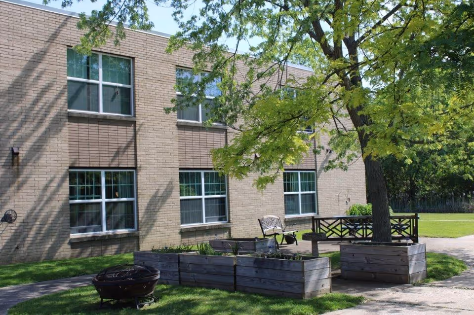 Outdoor area of a senior living facility with a brick building in the background, several windows, a large tree with green leaves, wooden planter boxes, a metal fire pit, and benches on a grassy lawn under a sunny sky.