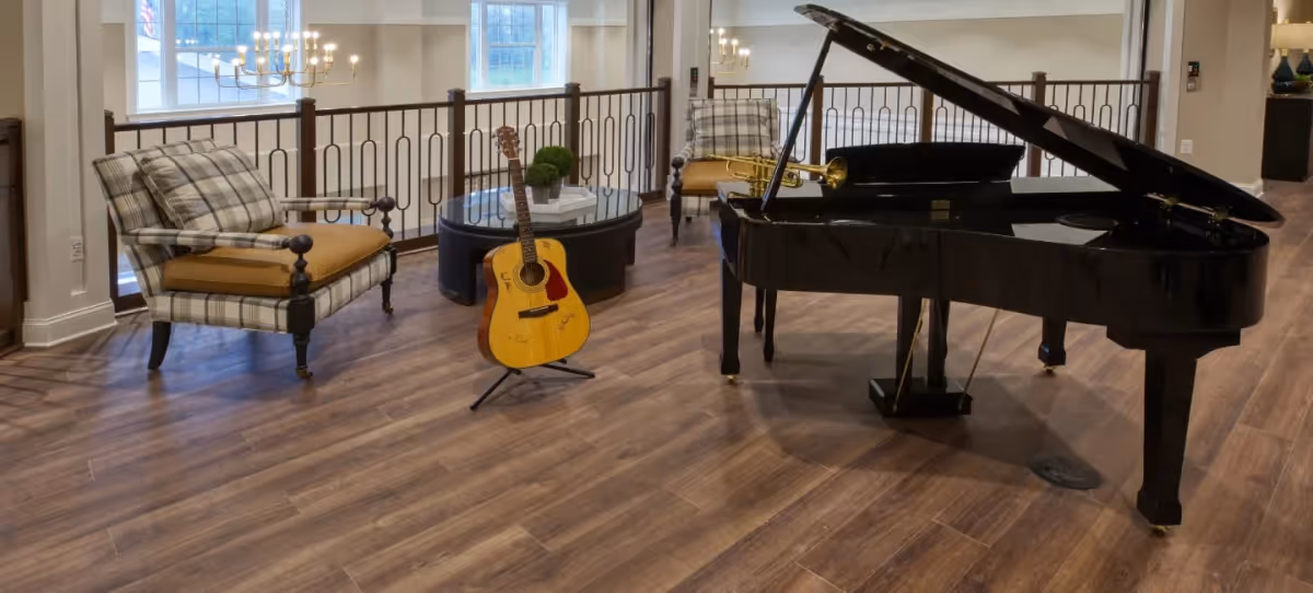 A cozy interior lounge area featuring a black grand piano with its lid open, a yellow acoustic guitar on a stand, and two plaid armchairs with mustard cushions. A round glass coffee table with decorative greenery is positioned between the chairs. The room has wooden flooring and large windows letting in natural light.