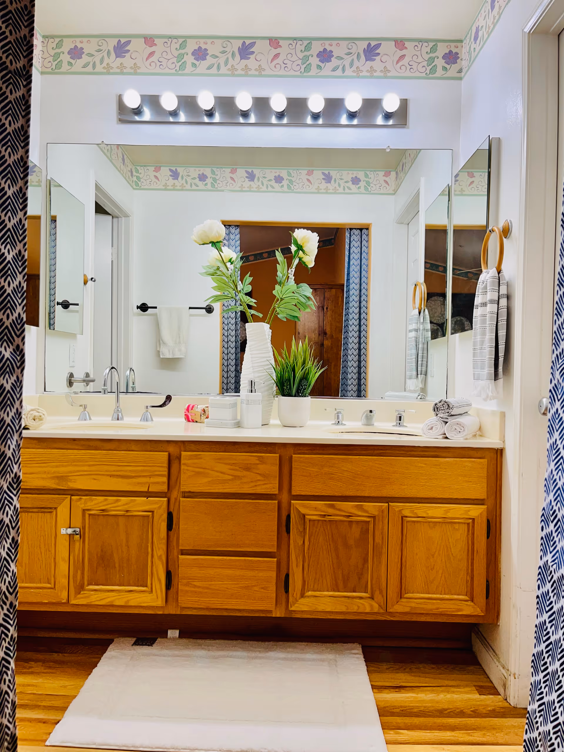 Bathroom vanity with double sinks, wooden cabinets, a large mirror, lit vanity bulbs, decorative plants and towels.
