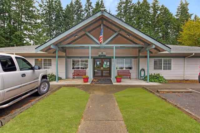 Front entrance of a single-story memory care building with a covered gabled porch, American flag, benches, potted plants, and parked vehicles.