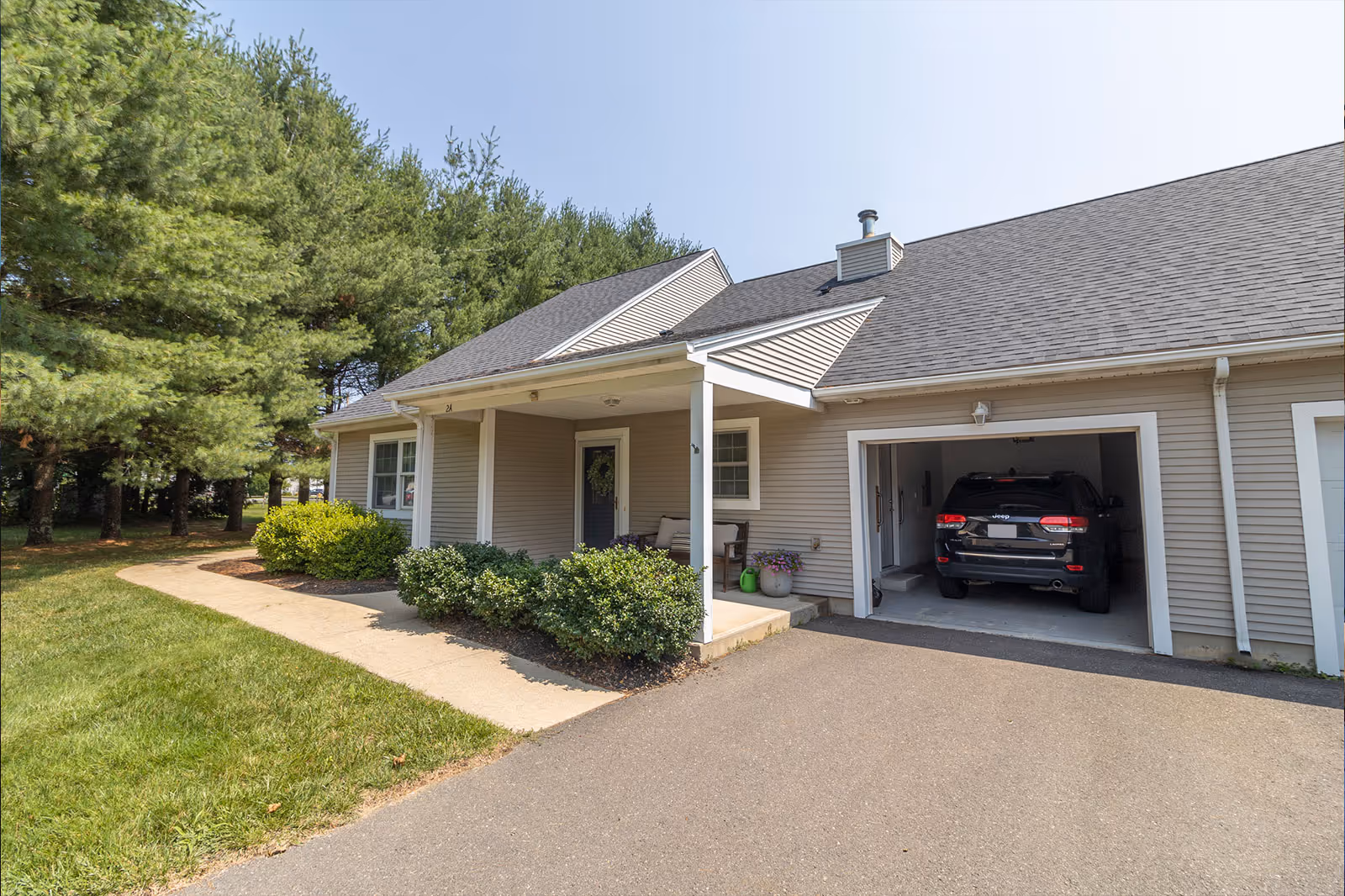 Single-story residential building with a covered front porch, attached garage with a parked SUV, driveway and landscaped shrubs.