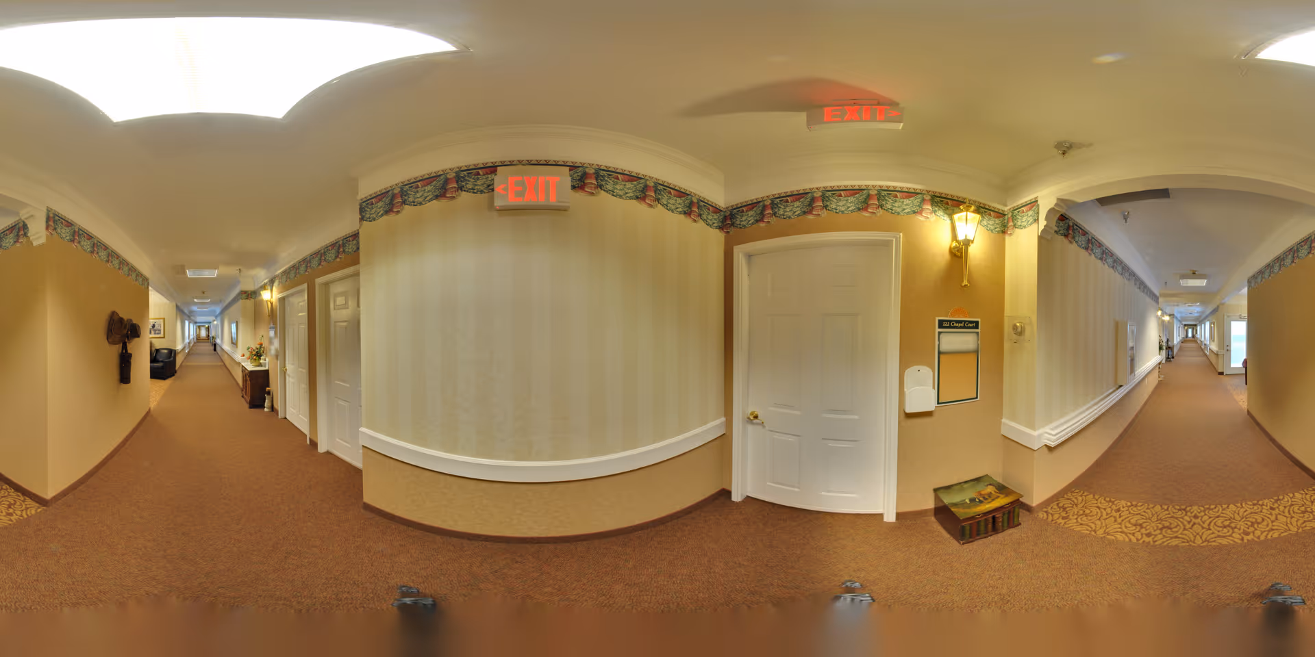 A long, carpeted hallway in a senior living facility with beige walls, white doors, and decorative wallpaper border near the ceiling. There are exit signs above the doors, wall-mounted light fixtures, and a small wooden bench with a cushion. The hallway extends into the distance with handrails along the walls.