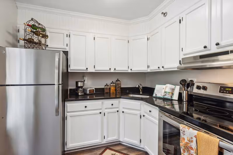 A modern kitchen with white cabinets, black countertops, a stainless steel refrigerator, an electric stove with a digital clock, a coffee maker, three glass jars with food items, a cookbook on a stand, and kitchen utensils in a holder.