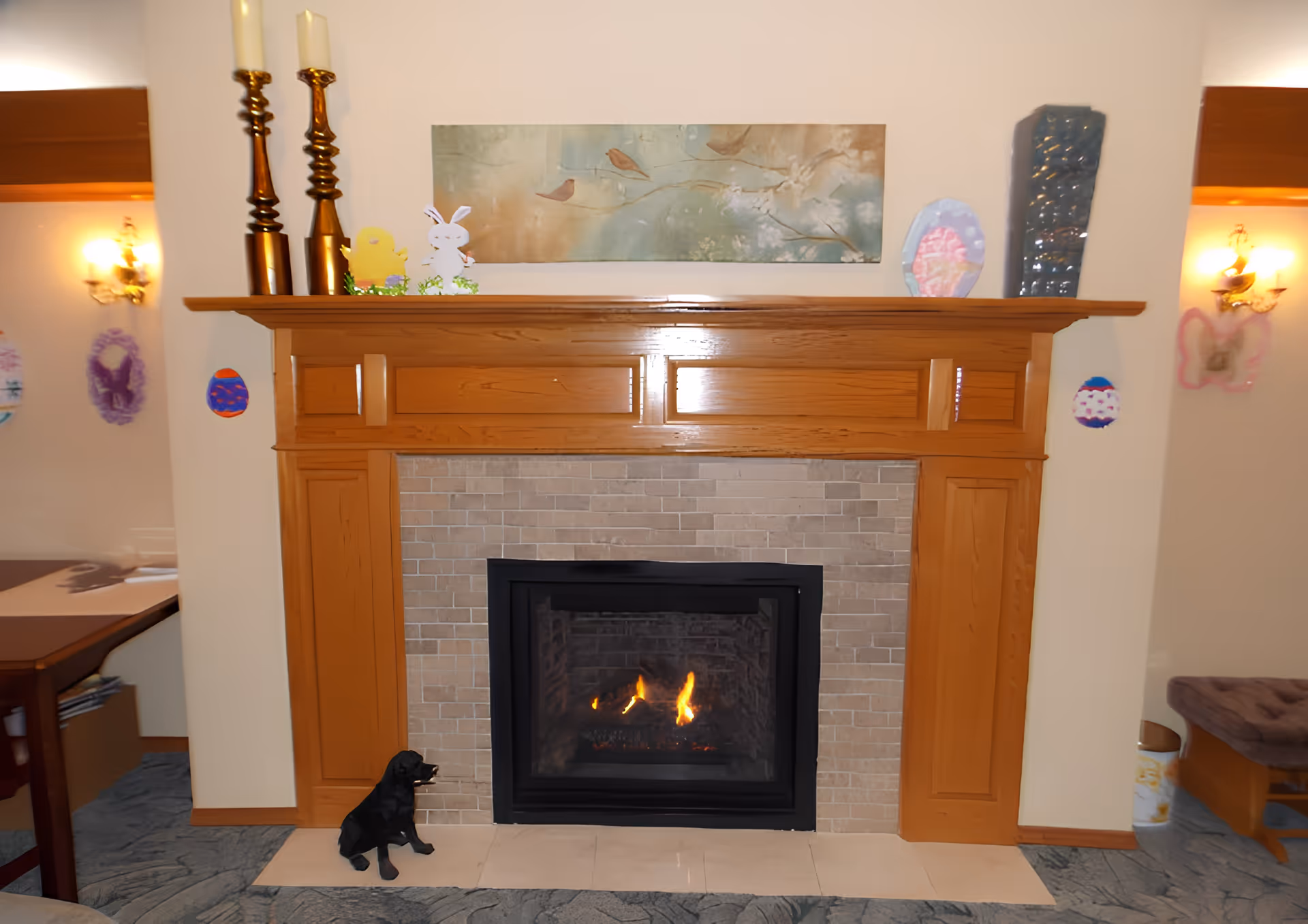 Lit fireplace with a wooden mantel decorated with candles, artwork, and Easter decorations in a communal living area.