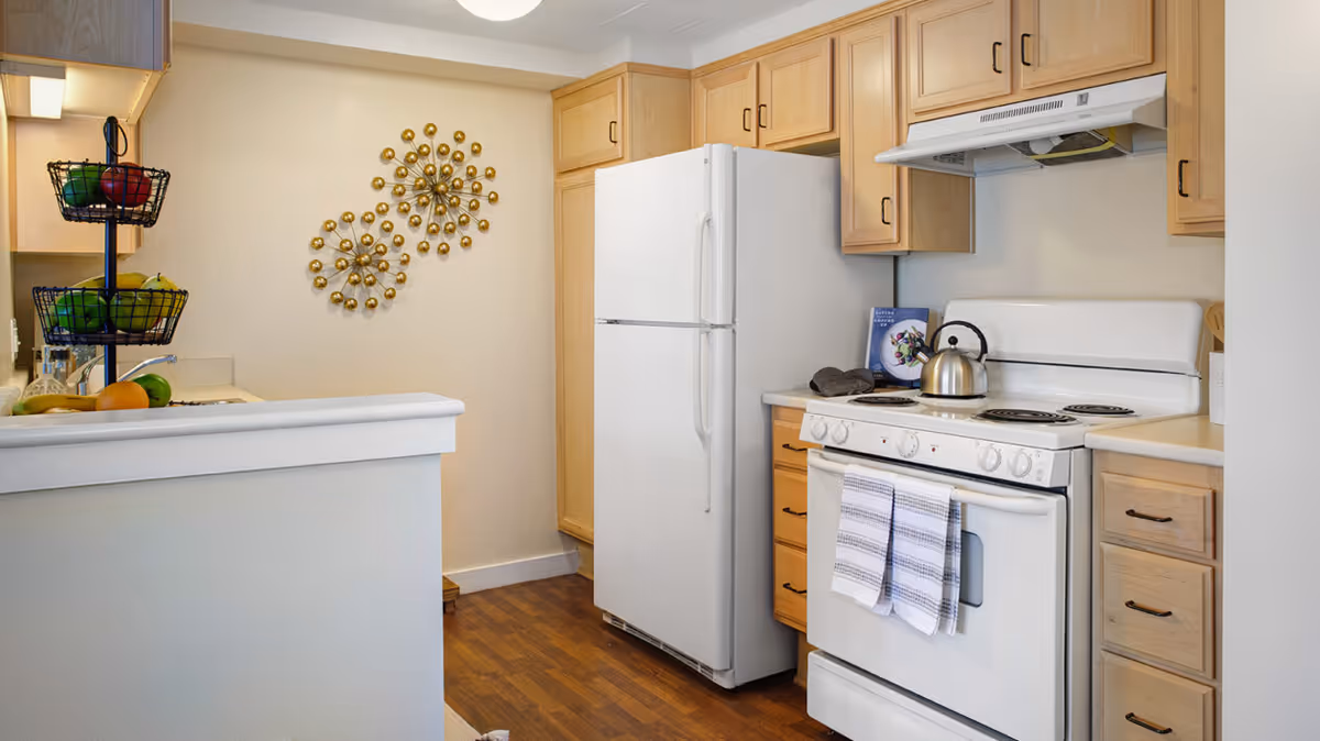 A compact kitchen with light wood cabinets, a white refrigerator, and a white electric stove with a kettle on top. Two striped kitchen towels hang on the oven door. A three-tiered fruit basket with various fruits is on the counter. The wall features two decorative gold metal wall art pieces. The floor is wood, and the walls are painted light beige.