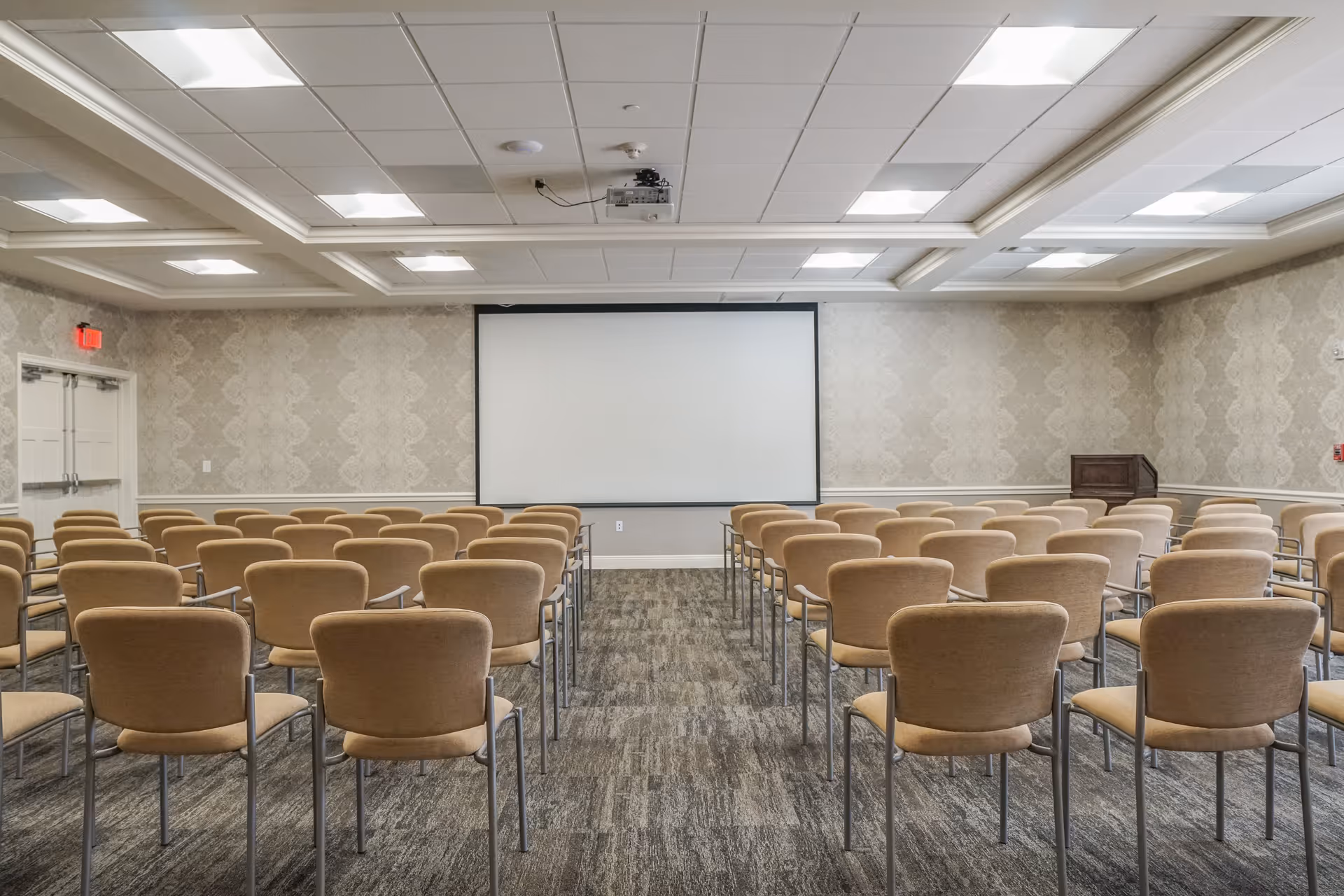 A conference or meeting room with rows of beige chairs facing a large white projection screen mounted on a patterned wallpapered wall. The room has a carpeted floor, a ceiling-mounted projector, and a wooden podium to the right side of the screen. Double doors are visible on the left side of the room.