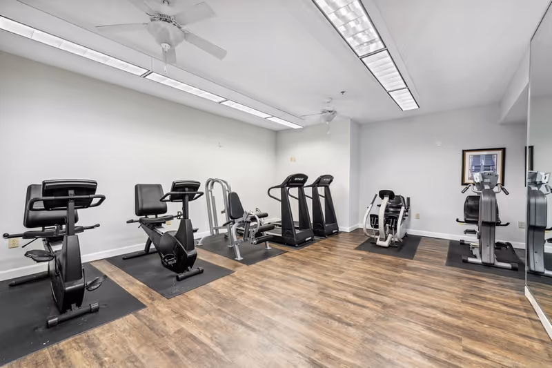 A fitness room with exercise equipment including stationary bikes, treadmills, and rowing machines on black mats. The room has wood flooring, white walls, ceiling fans, and fluorescent lighting. A large mirror covers part of the right wall.