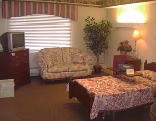 A furnished senior bedroom featuring a single bed with a floral bedspread, a matching floral couch, a TV on a dresser, and a lamp beside a potted plant.