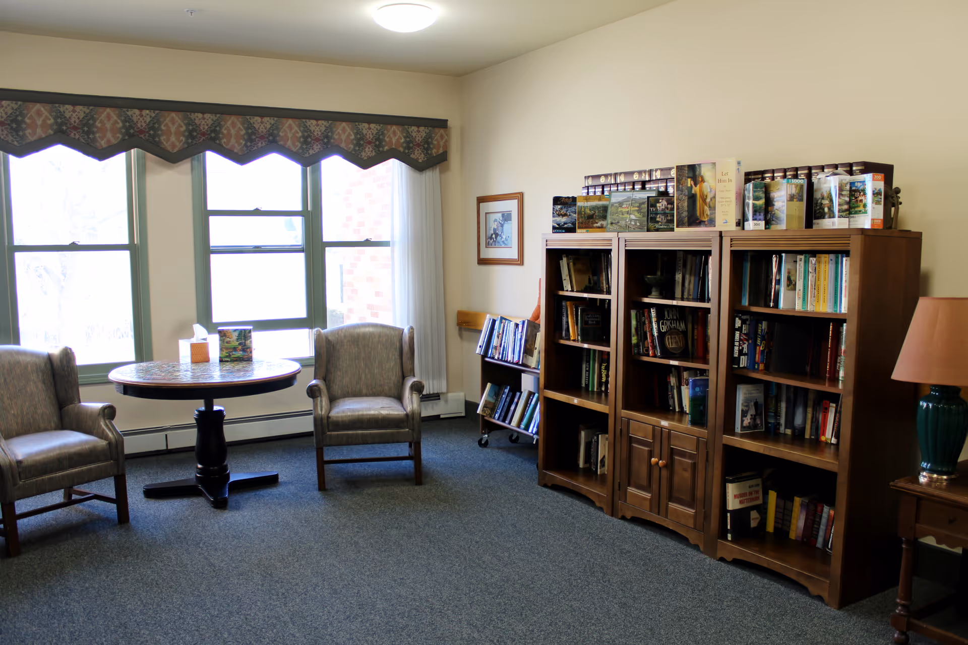 A cozy reading or sitting area with two upholstered armchairs around a small round table near three large windows with patterned valances. There are three wooden bookshelves filled with books and some decorative items on top. A small lamp with a green base and a pink shade sits on a side table to the right. The room has beige walls and a blue carpeted floor.