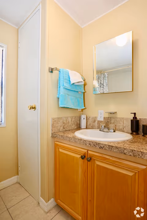 Bathroom sink area with a wooden cabinet below, a countertop with a built-in sink, a soap dispenser, and a small bottle. Above the sink is a rectangular mirror. To the left, a towel rack holds folded blue and white towels. A closed white door is visible next to the towel rack.
