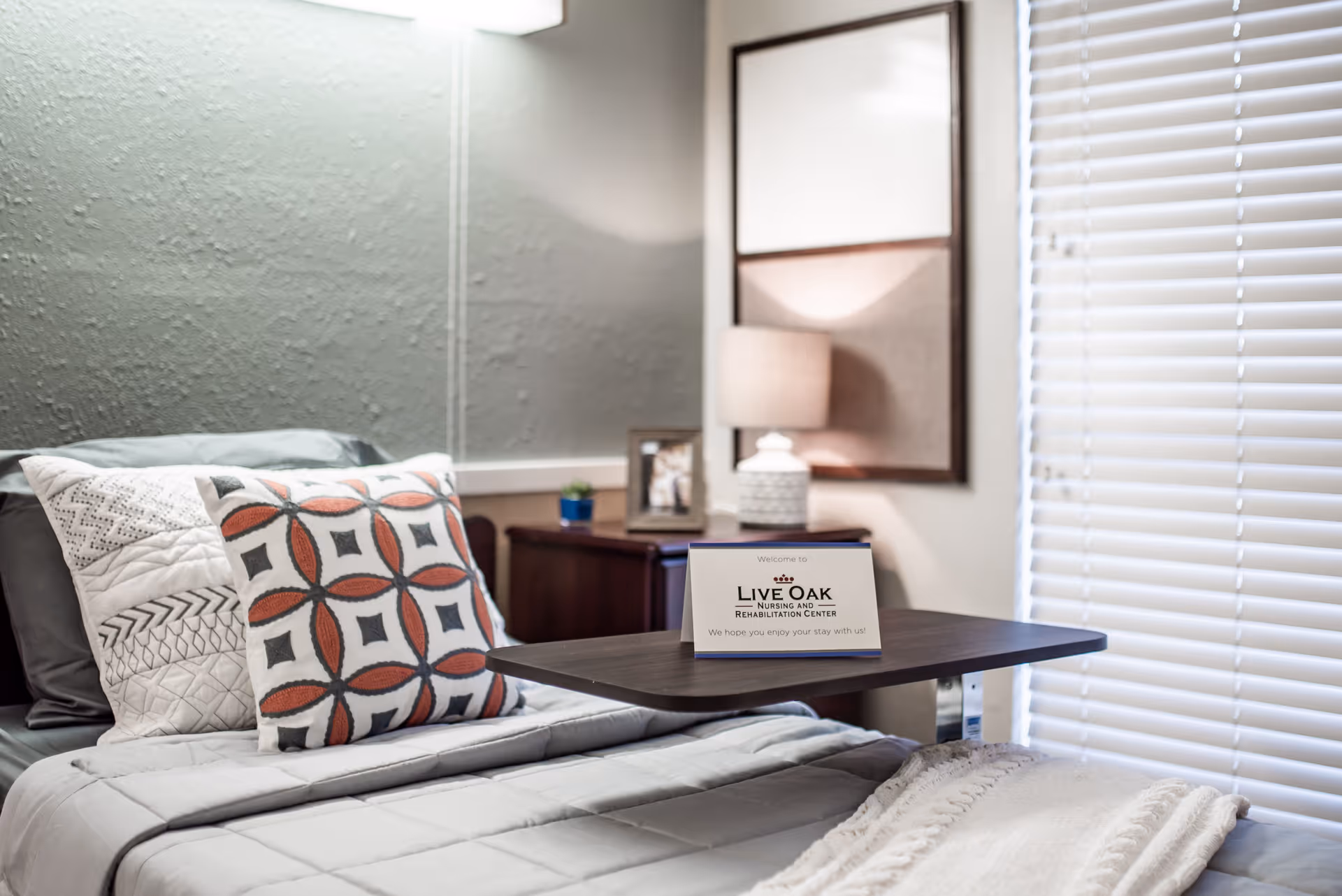 A neatly made bed with decorative pillows and a gray quilted blanket in a nursing and rehabilitation center room. A wooden overbed table holds a welcome sign for Live Oak Nursing and Rehabilitation Center. In the background, there is a nightstand with a lamp, a small plant, and a framed photo, next to a window with closed blinds.