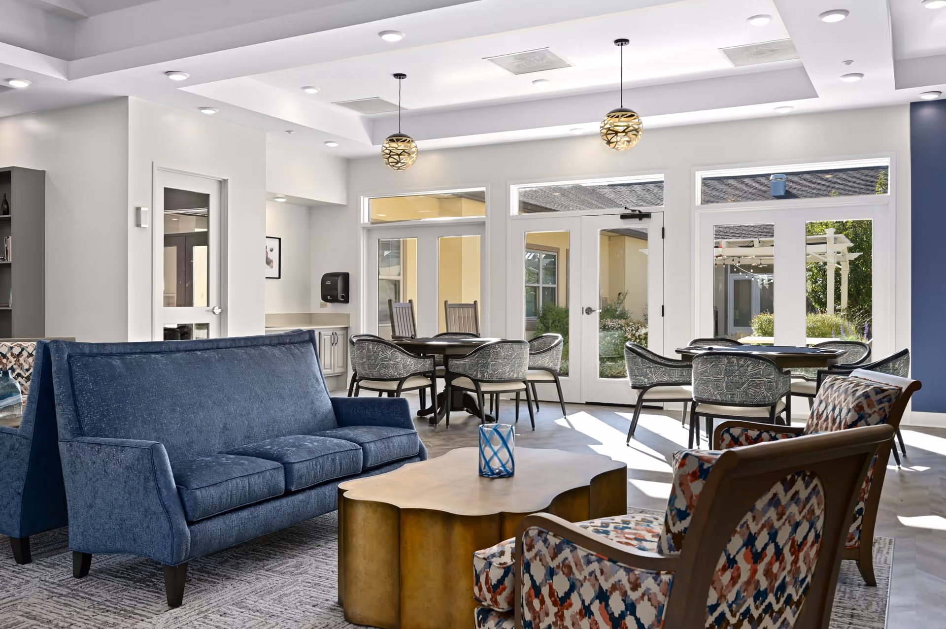 A bright and modern common area with blue upholstered sofas, patterned armchairs, a wooden coffee table, and round tables with chairs near large windows and glass doors leading to an outdoor space.