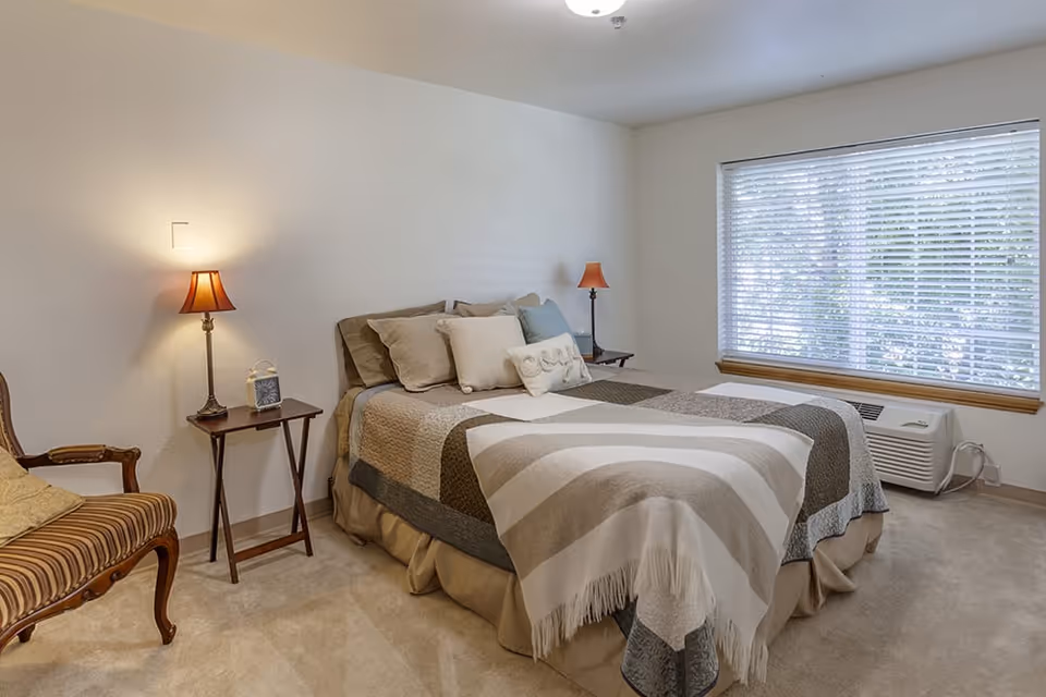 A neatly made bedroom with a bed, bedside tables and lamps, a striped armchair, and a window with blinds.