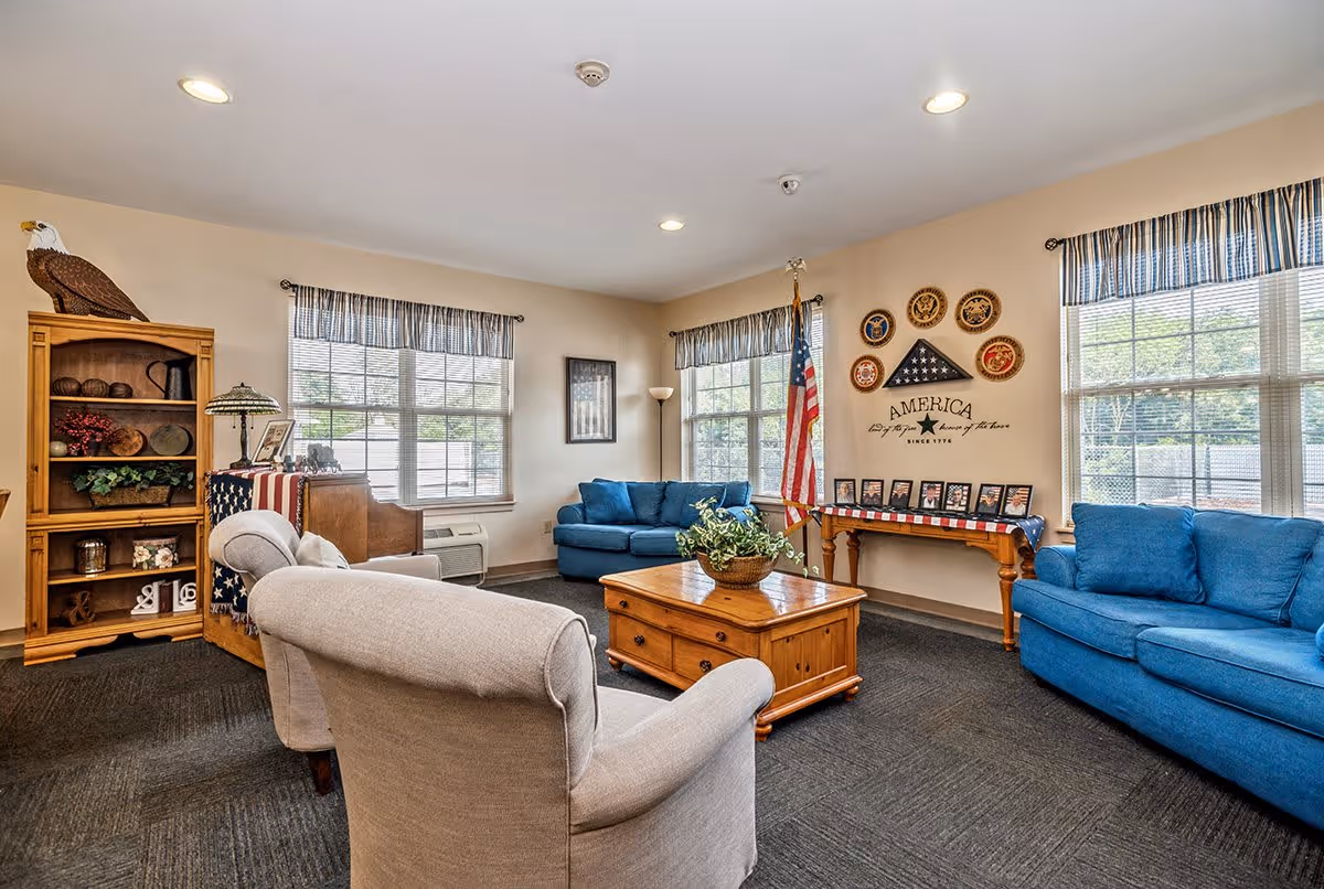 A cozy living room with beige walls and carpeted floor featuring two blue sofas and two beige armchairs arranged around a wooden coffee table with a plant centerpiece. The room has three windows with striped valances allowing natural light to enter. On one wall, there is an American flag, patriotic wall decor including military insignias, and a table draped with an American flag displaying framed photos. A wooden bookshelf with decorative items and a lamp is positioned in the corner.