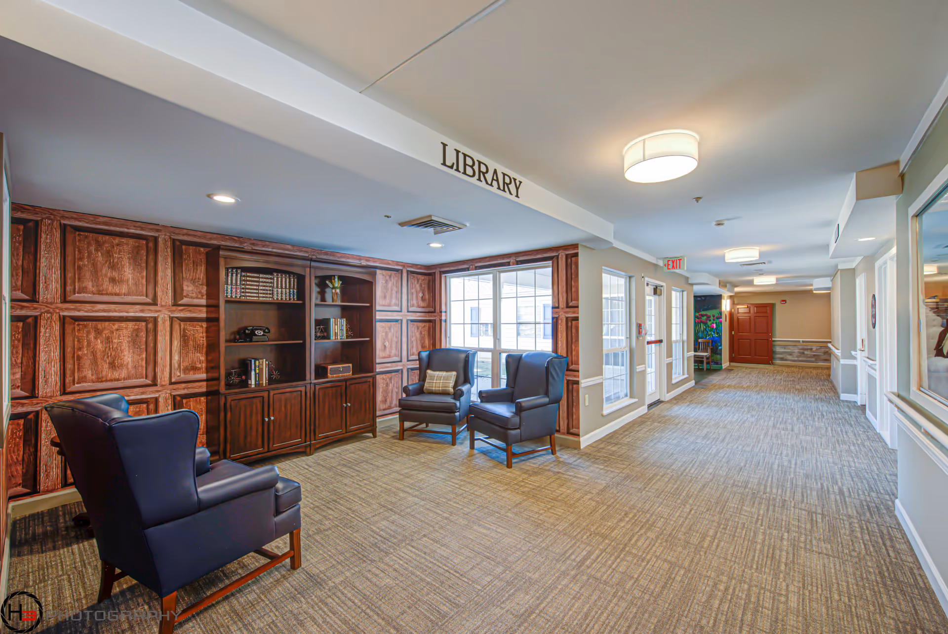Interior view of a library area in a senior living facility with three dark blue armchairs arranged near a wooden bookshelf filled with books and decorative items. The walls have wood paneling, and large windows allow natural light to enter. The carpeted hallway extends to the right with ceiling lights and an exit sign visible.