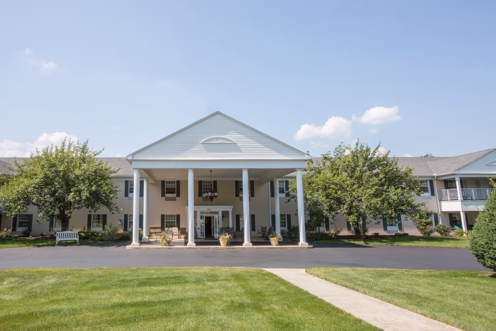 Front exterior view of a senior living facility named Country Meadows of Bethlehem with a large covered entrance supported by white columns, surrounded by green trees and a well-maintained lawn under a blue sky with a few clouds.