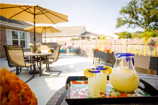 Outdoor patio area with a table holding a tray containing a pitcher of lemonade and two glasses garnished with lemon slices. In the background, there are patio chairs, tables with umbrellas, and a wooden fence with flower planters.