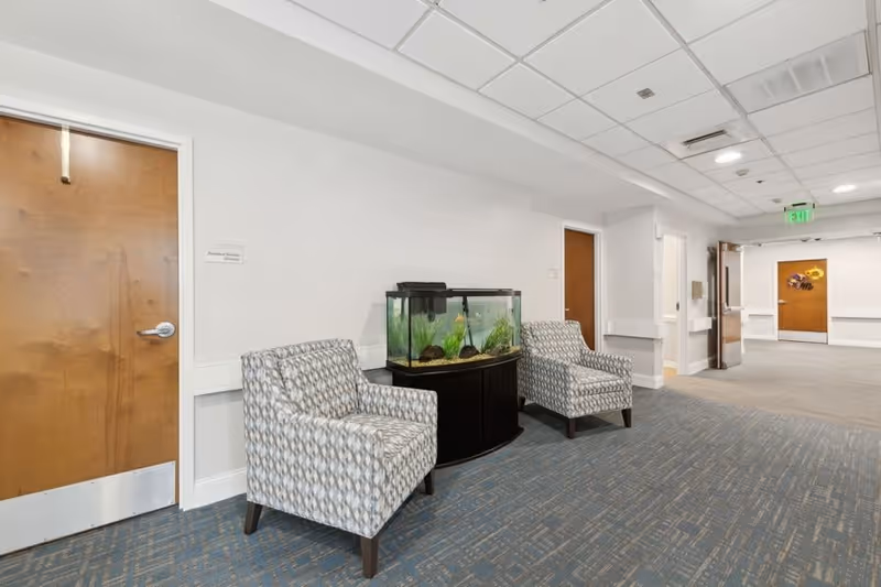 A hallway in a senior living facility with two patterned armchairs placed on either side of a fish tank on a black stand. The hallway has white walls, a blue patterned carpet, wooden doors, and ceiling lights. An exit sign is visible in the background.
