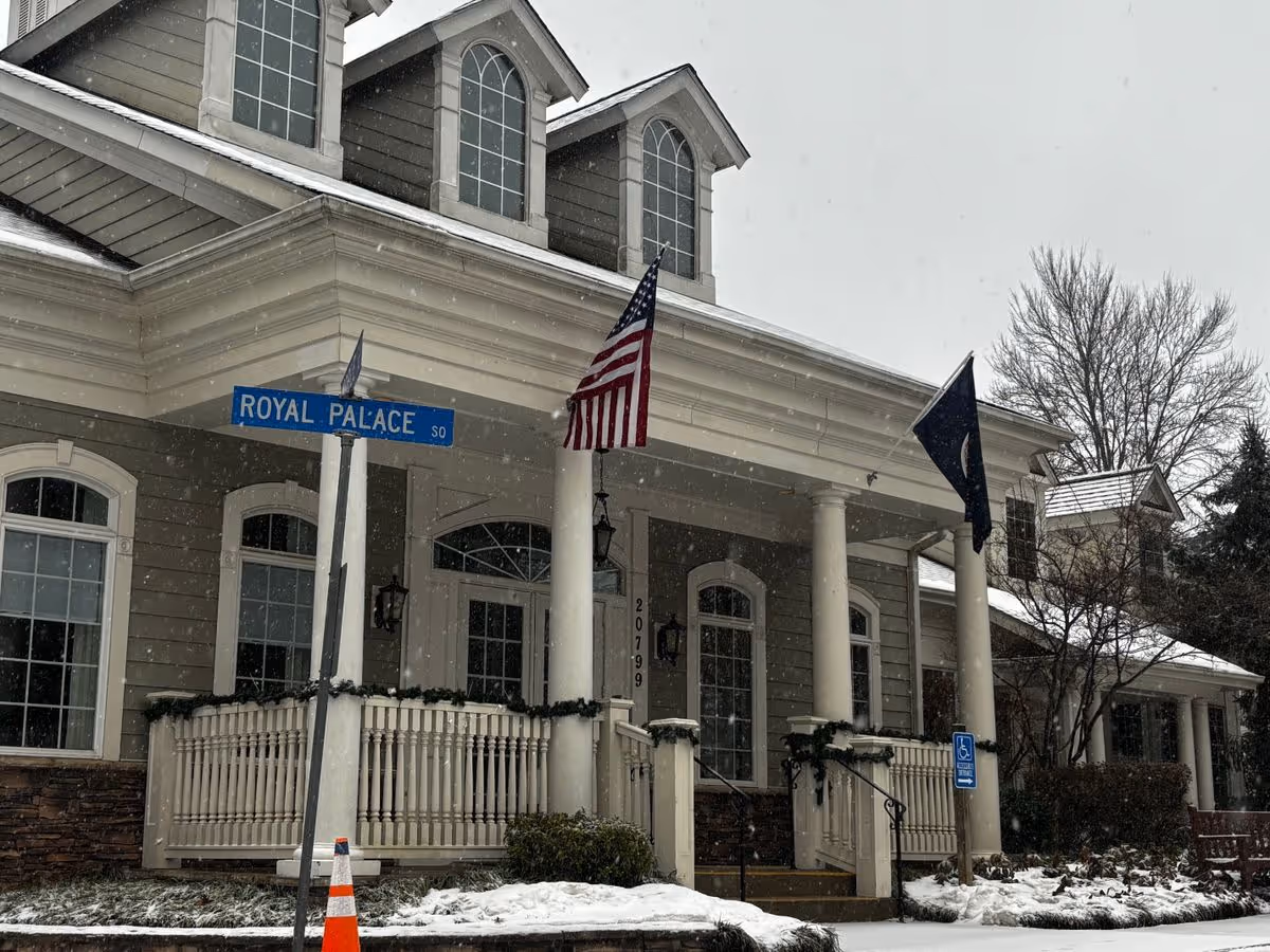 Snow falling at the front entrance of a Colonial-style building with columns, an American flag, and a 'Royal Palace' street sign.