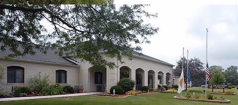 Front exterior of a single-story brick senior living building with arched entrances, landscaped lawn, and flagpoles.