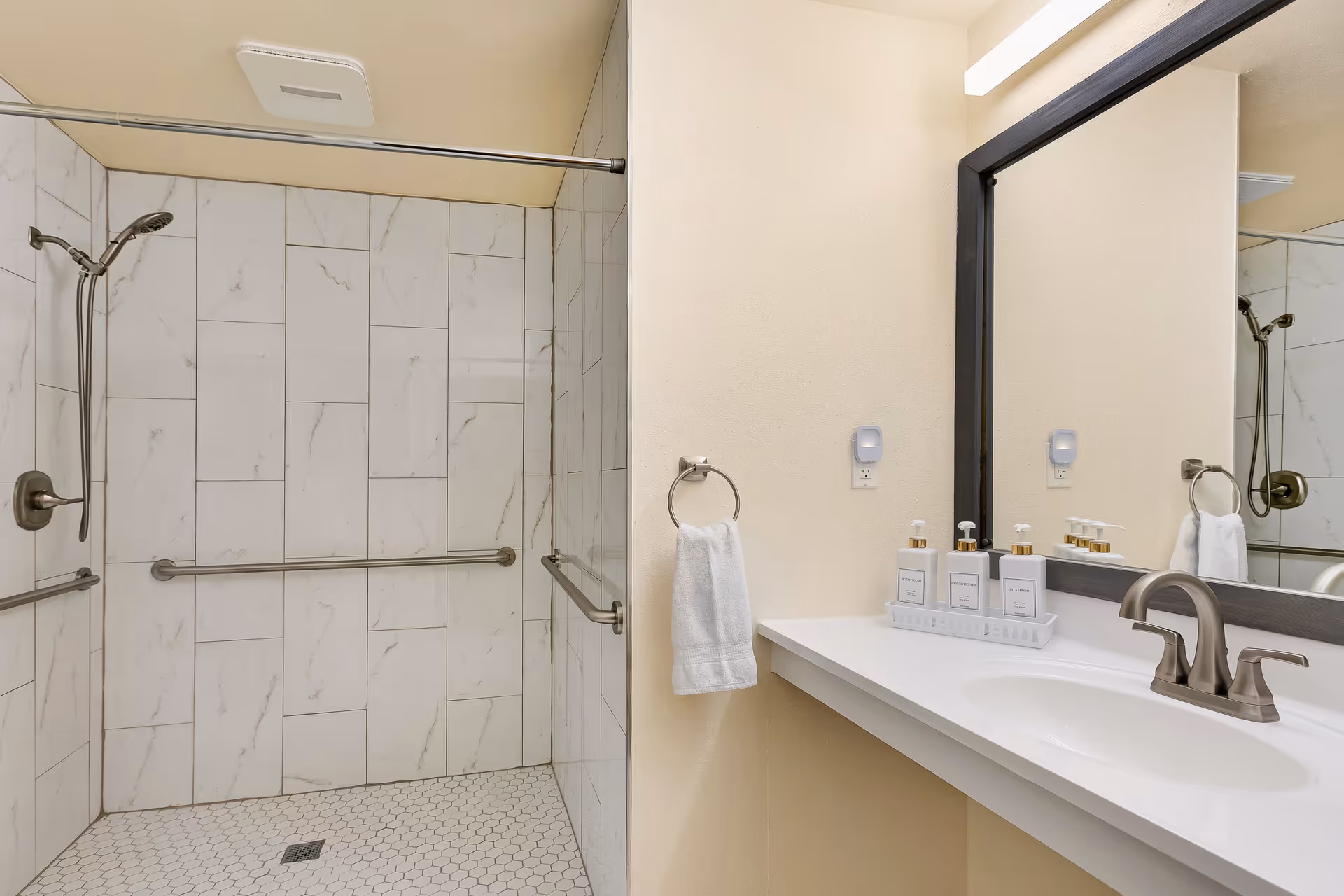 A clean and modern bathroom featuring a walk-in shower with white marble-patterned tiles and a handheld showerhead. The shower has stainless steel grab bars for safety. Adjacent to the shower is a white countertop with a built-in sink, a silver faucet, and three pump bottles labeled body wash, conditioner, and shampoo. A white towel hangs on a ring next to the sink, and a large mirror is mounted on the wall above the countertop.