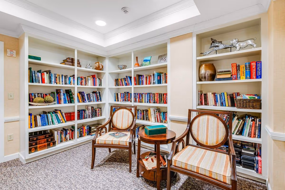 A cozy reading nook in a senior living facility featuring built-in white bookshelves filled with books and decorative items. Two wooden armchairs with striped upholstery are positioned around a small round wooden table with a green box and some papers on it. The room has beige walls and carpeted flooring.