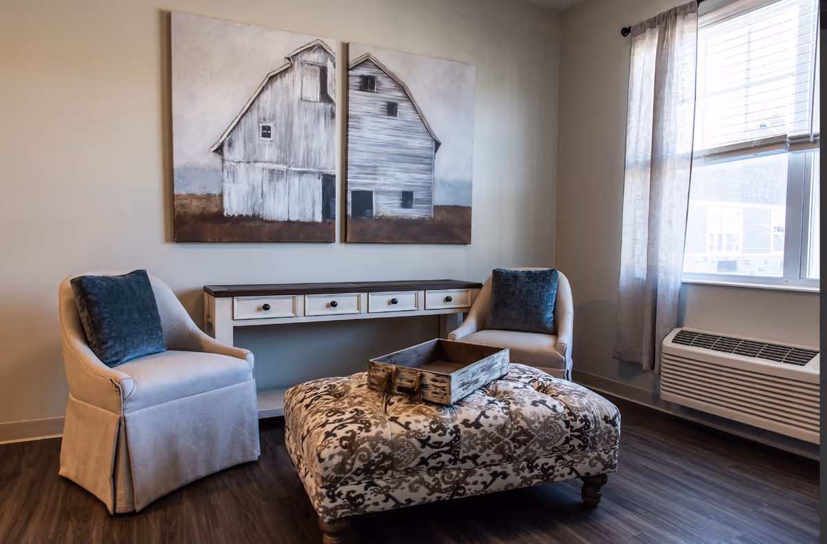 A cozy sitting area with two beige armchairs each with a dark blue cushion, a patterned ottoman with a rustic wooden tray on top, a narrow console table with drawers against the wall, and two large paintings of barns hanging above the table. A window with sheer curtains allows natural light into the room.