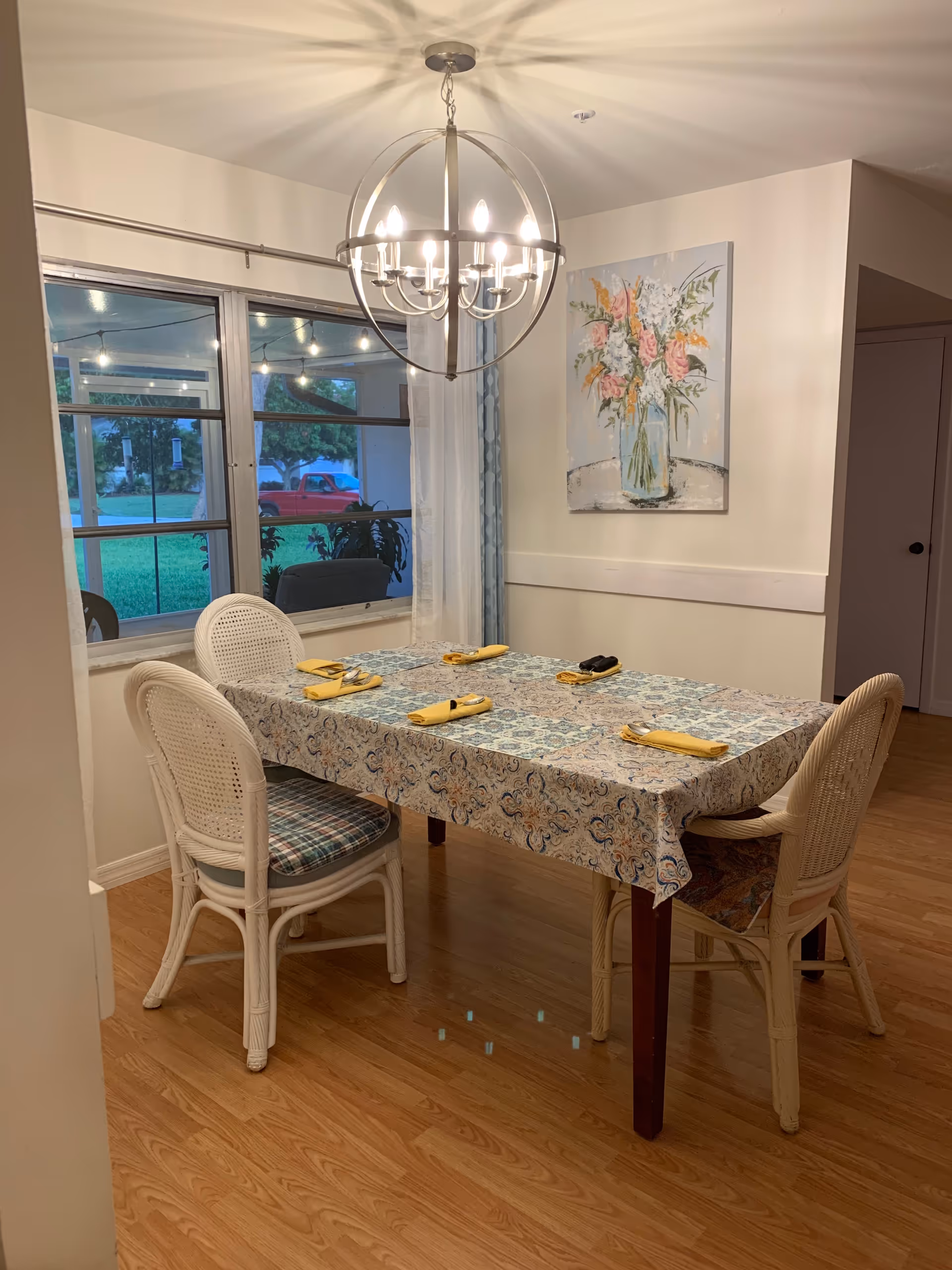 A dining area with a rectangular table covered with a patterned tablecloth. Four wicker chairs with cushions surround the table, which is set with yellow napkins and silverware. A modern chandelier with multiple lights hangs above the table. A large window with sheer curtains shows an outdoor view with string lights and greenery. A floral painting is hung on the wall to the right.