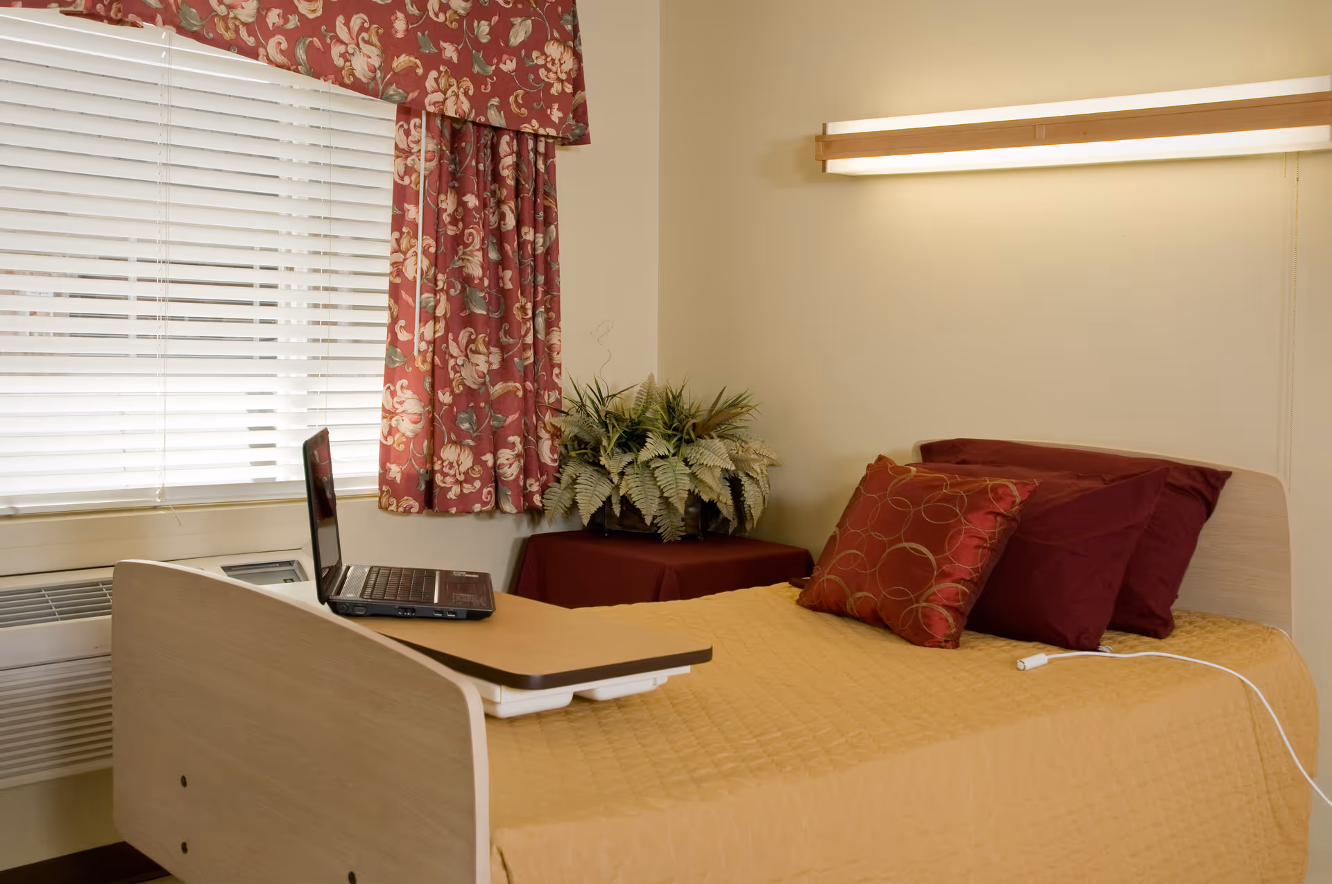 A neatly made single bed in a residential room with a laptop on an over-bed table, red patterned curtains, decorative pillows, and a potted plant.