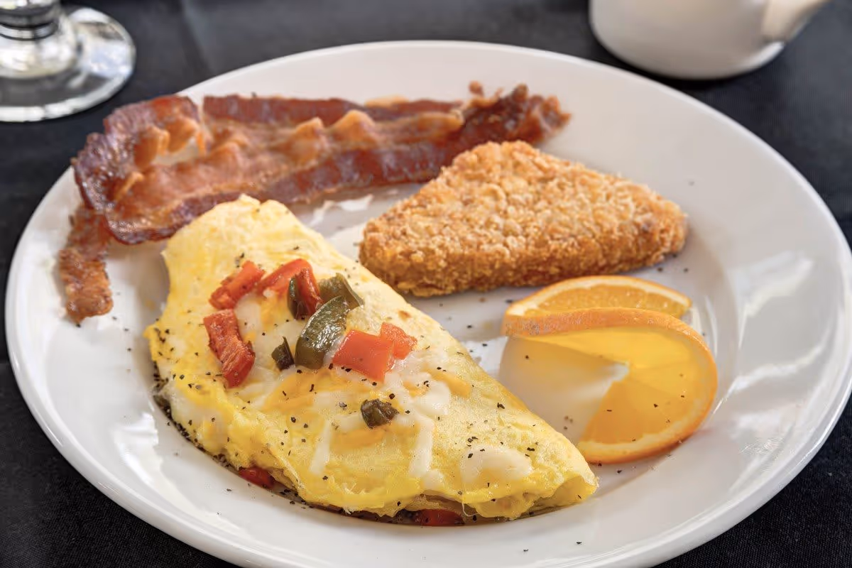 A breakfast plate with an omelette topped with diced tomatoes and green peppers, two strips of crispy bacon, a triangular hash brown, and two orange slices on a white plate with a black tablecloth underneath.