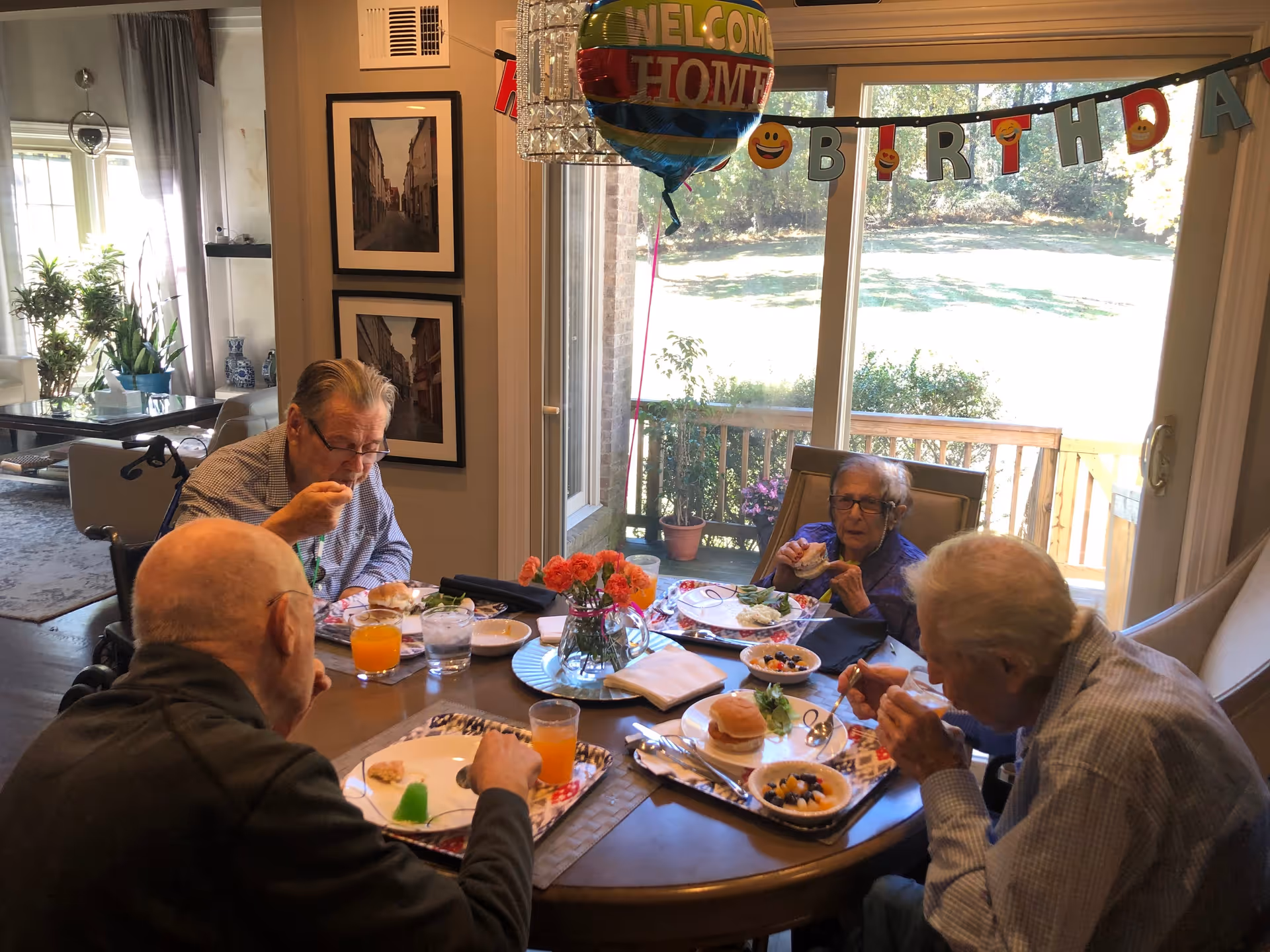 Four elderly people sitting around a dining table enjoying a meal together in a well-lit room with large windows showing a garden outside. There is a 'Welcome Home' balloon and a 'Happy Birthday' banner hanging near the window. The table has plates with food, glasses of juice, and a vase with flowers in the center.