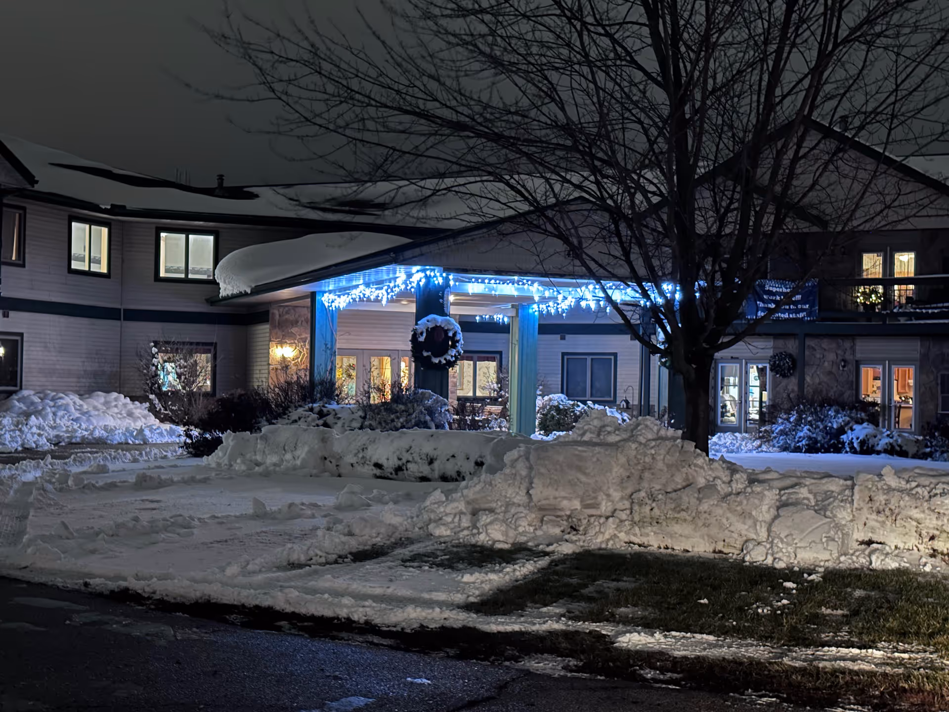 Nighttime exterior view of Stonefield Apartment Homes & Health Services building entrance decorated with blue holiday lights and a wreath. Snow covers the ground and roof, with a leafless tree in front of the entrance.