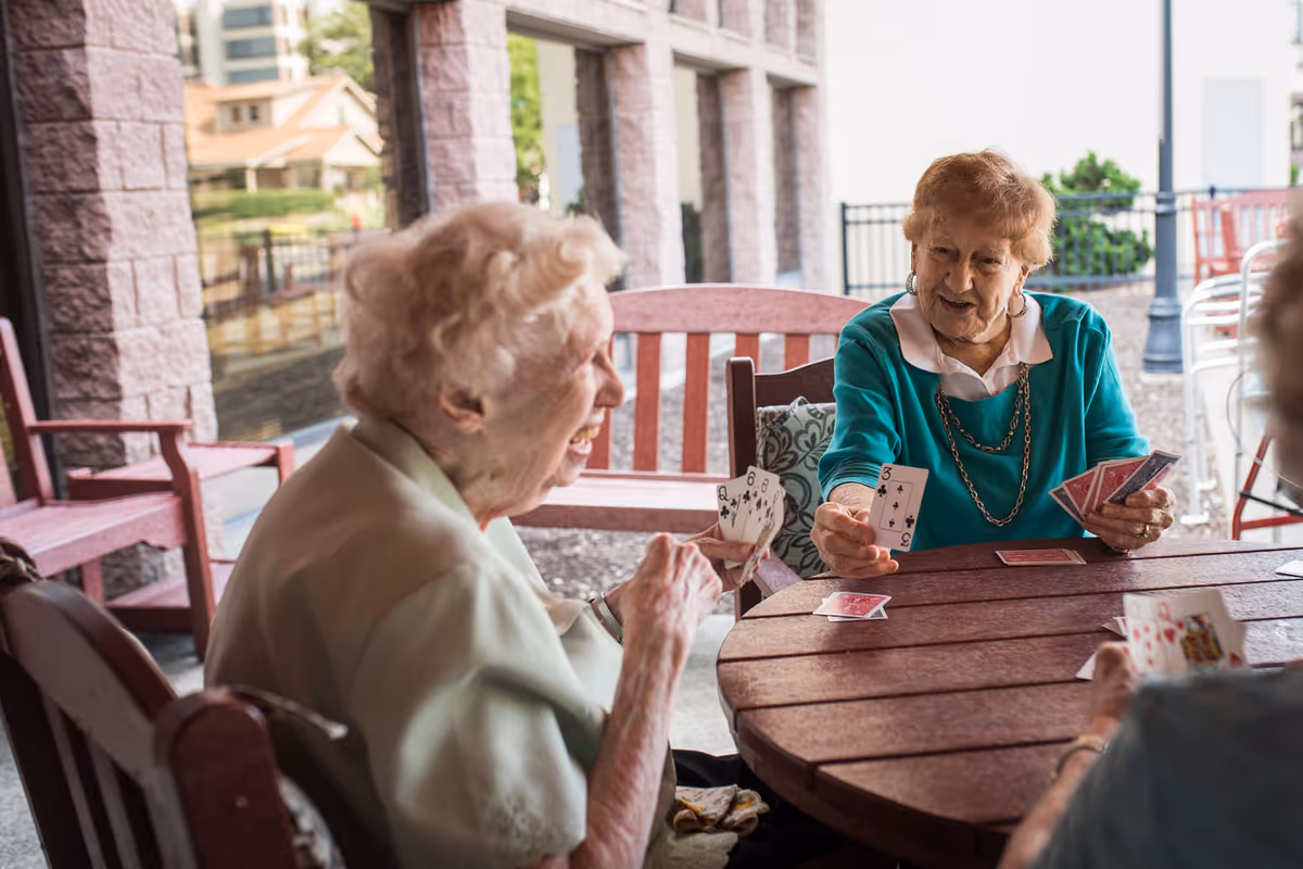 Three elderly women sitting around a round wooden table on a covered patio, playing a card game and smiling. The background shows stone pillars and outdoor furniture with a view of nearby buildings and greenery.