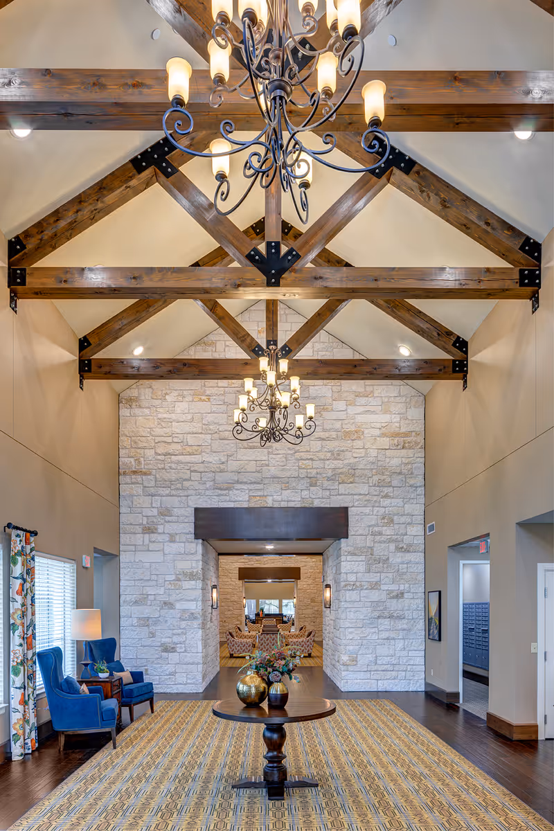 Interior view of a senior living facility lobby with high vaulted ceiling featuring exposed wooden beams and two ornate chandeliers. A round wooden table with decorative vases and flowers is centered on a patterned carpet. To the left, there are two blue upholstered chairs next to a window with floral curtains and a floor lamp. The far wall is made of light-colored stone with a large opening leading to another seating area with armchairs and a mirror.