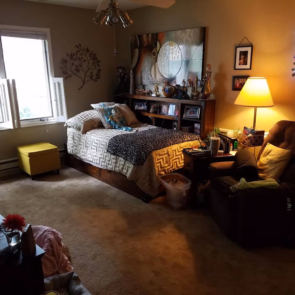 A cozy bedroom with a single bed covered in patterned bedding and a leopard print throw. The bed has several pillows, including one with a doll on it. Behind the bed is a wooden headboard with shelves displaying various decorative items and framed photos. To the right of the bed is a brown armchair with a yellow pillow and a small side table holding a lamp, books, and other items. A window with white shutters is on the left wall, letting in natural light. The room has beige walls and carpeted floor, with additional wall decorations and framed pictures.