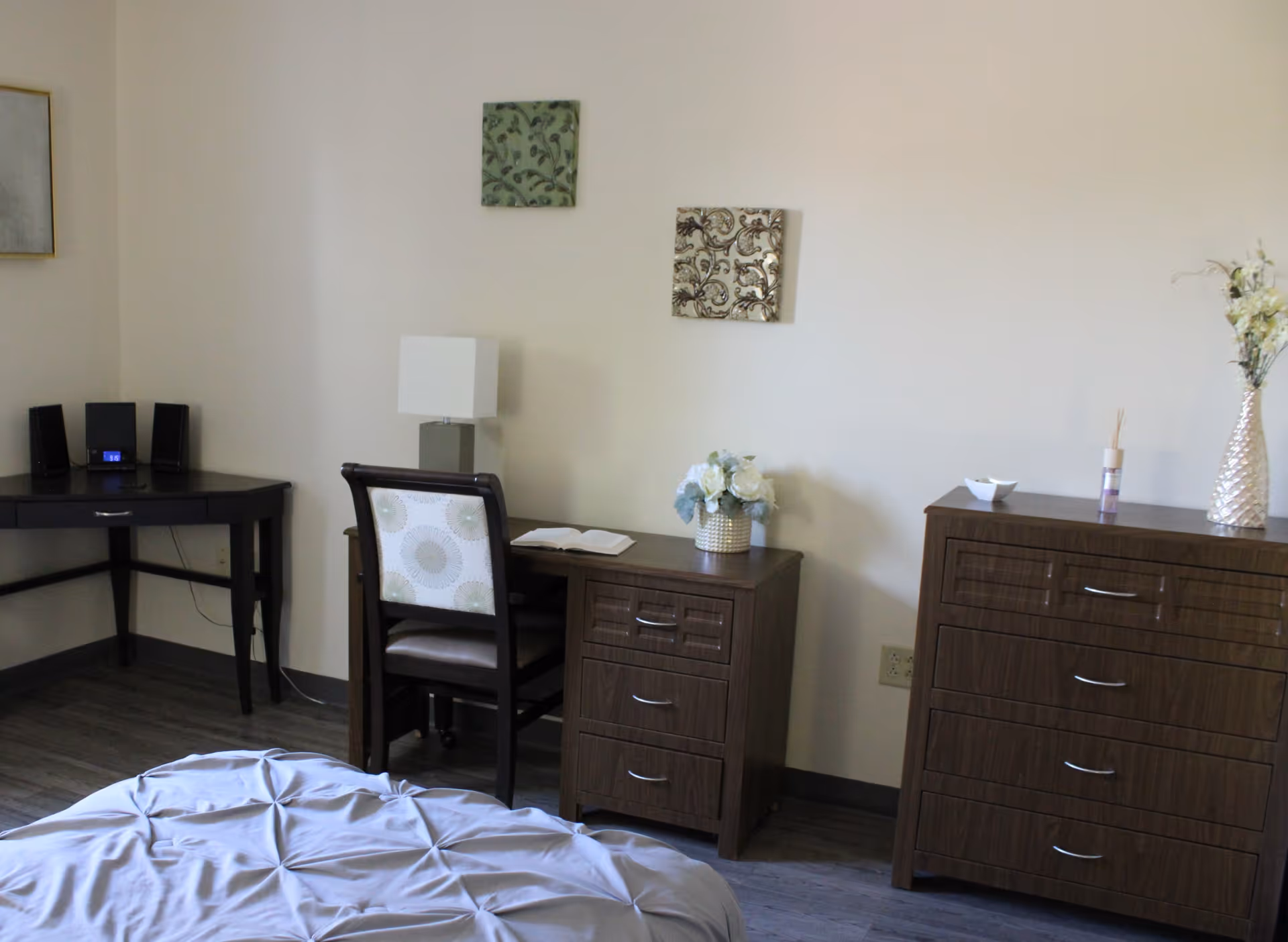 A bedroom corner featuring a wooden desk with a chair, a small lamp, and a vase with flowers. Next to the desk is a wooden dresser with multiple drawers, decorated with a vase of flowers and a small bowl. The bed with a light-colored comforter is partially visible in the foreground. The walls are light-colored with two small decorative wall hangings.