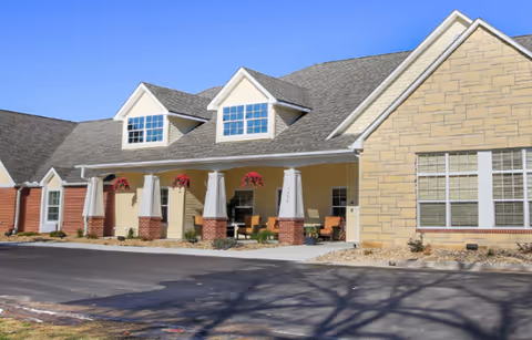 Front exterior of an assisted-living building showing a covered porch with columns, hanging flower baskets, seating, dormer windows, and a driveway.